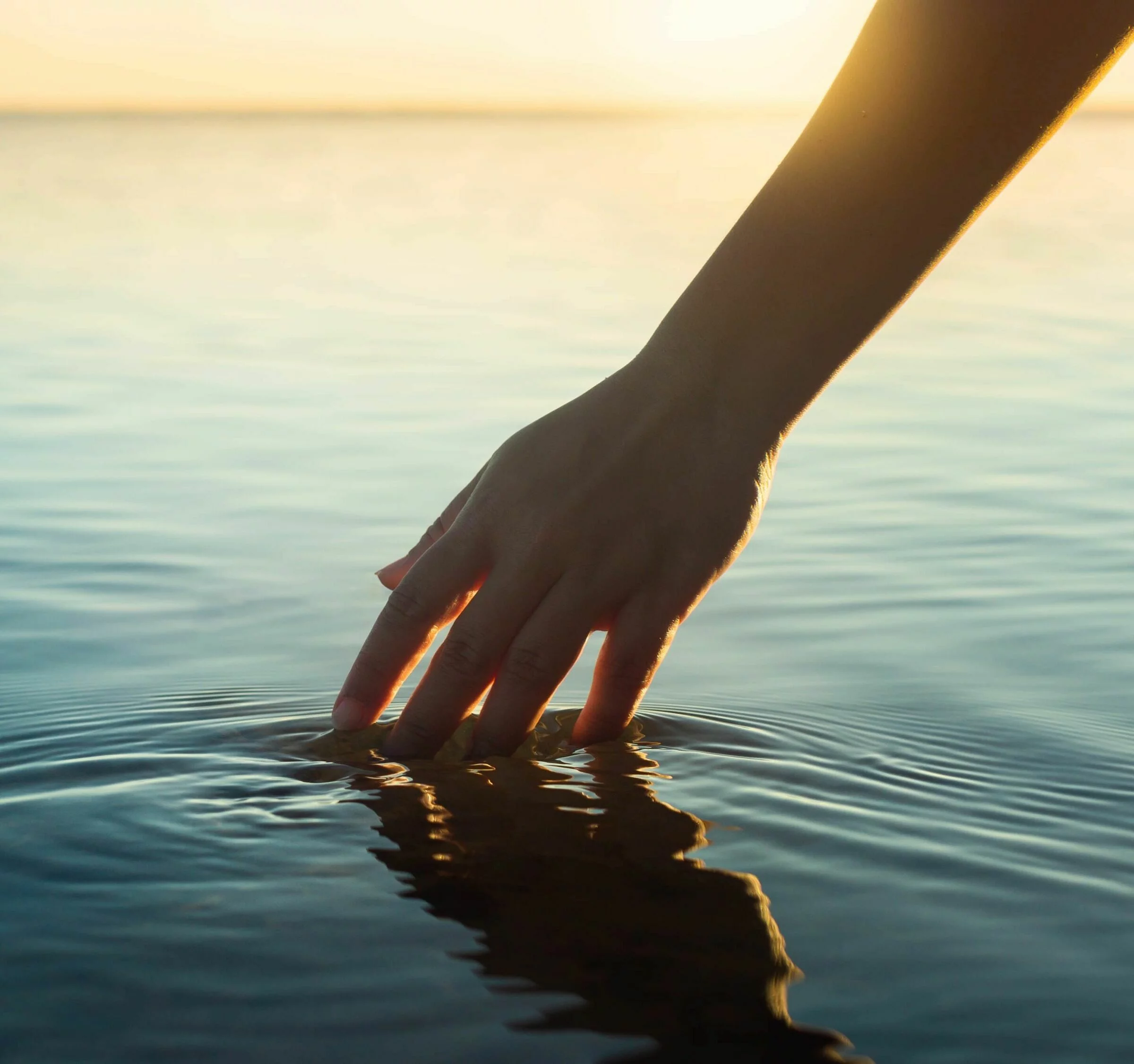 Menschenhand taucht in ruhiges Meerwasser bei Sonnenuntergang.